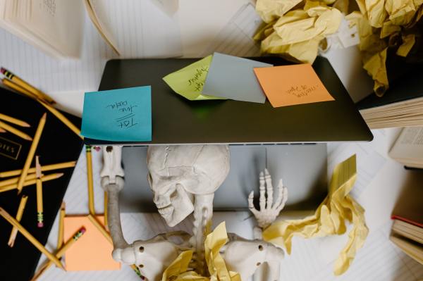 Ein Skelett liegt mit dem Kopf auf einem aufgeklappten Laptop. Um es herum liegen zerknüllte Papiere, zerbrochene Bleistifte, Haftnotizen und Bücher.